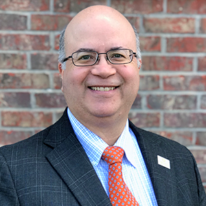 Octavio Martinez, photographed from the shoulders up. He stands against a brick background and wears a dark plaid patterned suit, light blue plaid button-up shirt underneath, and a red tie with blue spots. 