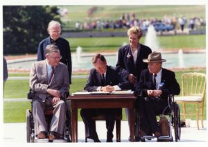 President H. W. Bush sits at a desk in the Rose Garden with two men using wheelchairs to sign the ADA