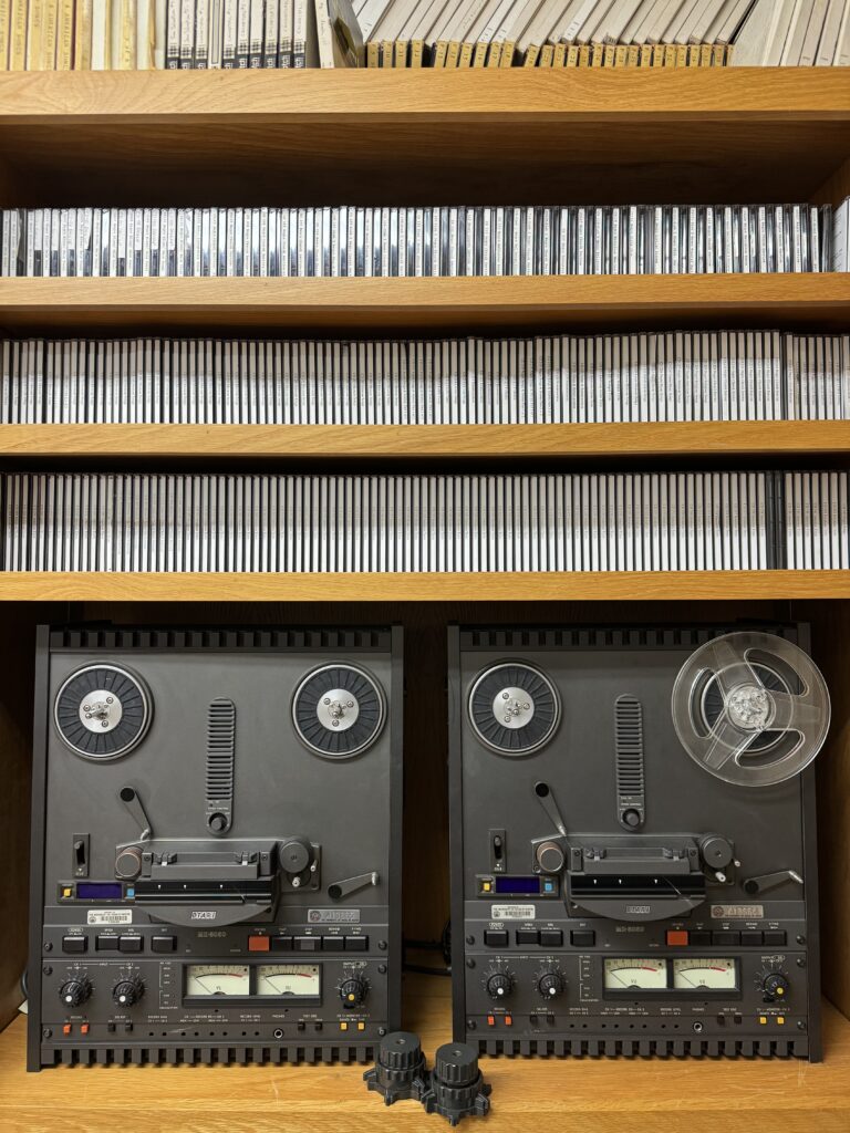 Photograph of two reel-to-reel audiotape machines with above them shelves of various types of recordings.