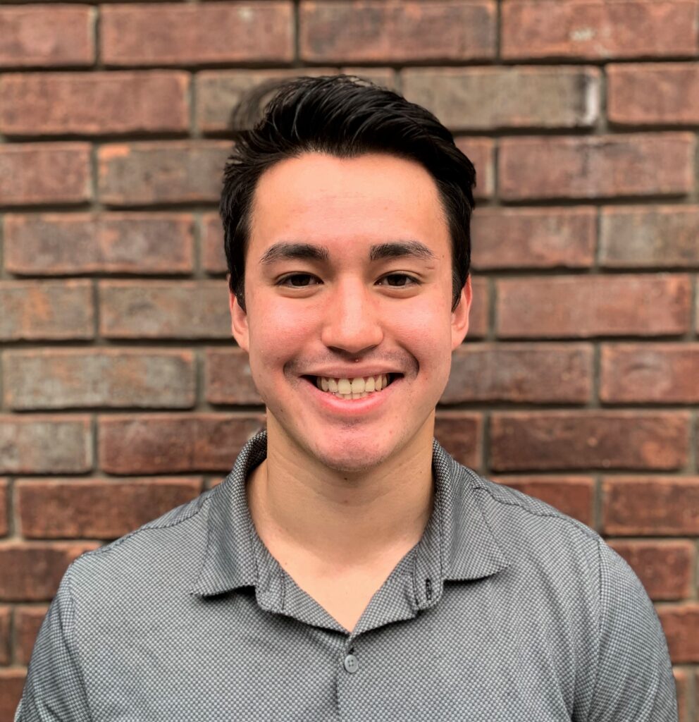 Colin Phillips is standing against a brick wall. He is wearing a grey collared shirt and is smiling.