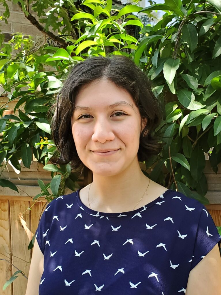 Vanessa Aguirre is standing in front of a tree wearing a navy top. She is smiling.