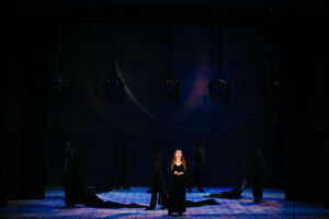 A woman sings on a darkly lit stage with ominous figures standing behind her clad all in black
