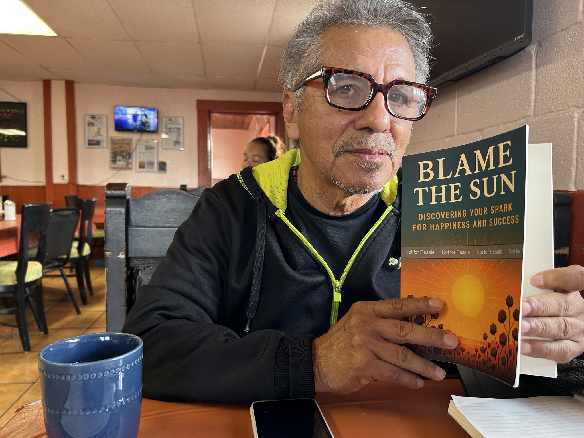 An elderly man sitting in a restaurant posing with a book called "Blame the Sun."
