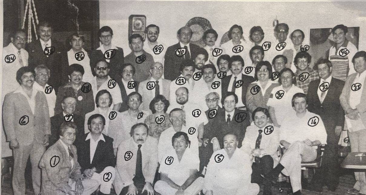 A large group of men and women sit and stand in a room, staring at the camera.