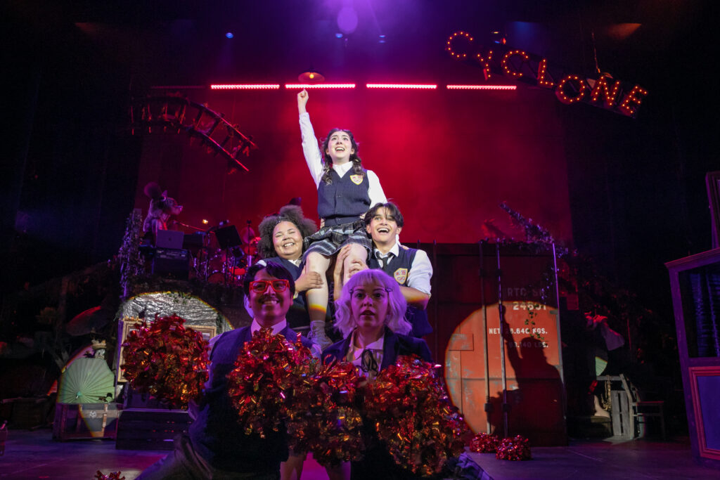 Students performing in the musical "Ride the Cyclone" - students are holding pom poms, and one student is raised up with her fist in the air