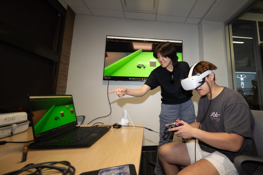 Student wearing a VR headset playing a VR game, with another student standing next to him helping guide him through the game