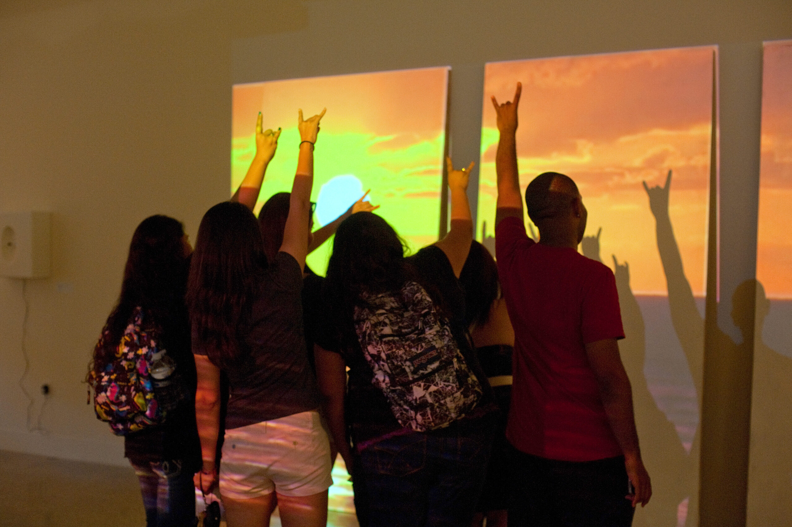 Six students stand in front of an art piece that is being projected onto a wall. They hold their hands up in a "hook 'em horns," casting shadows on the artwork on the wall.