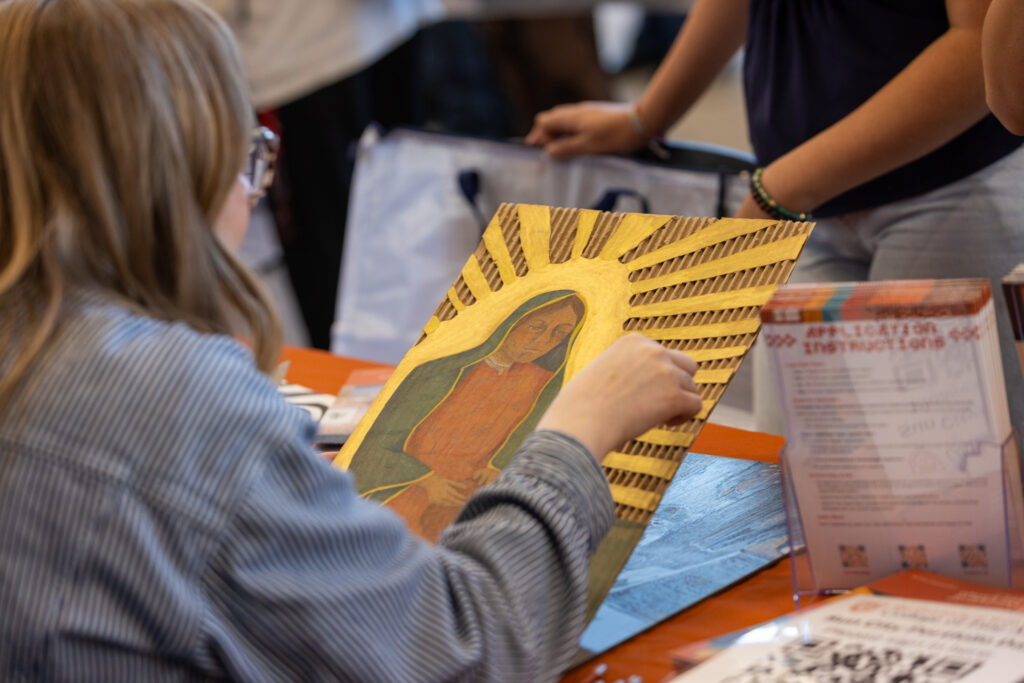 Some sits behind a table that has a UT table cloth and materials on it as they discuss a piece of art with the artist who created it. The art is made of cardboard and depicts a saint, with gold metallic rays coming out from behind the saint.
