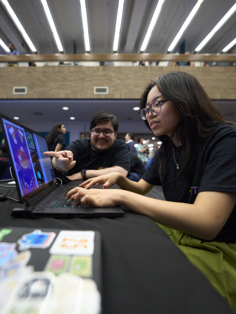 Two students sit at a table. One student is play testing a game on a laptop, while the other student points to the screen to help the student playing game.