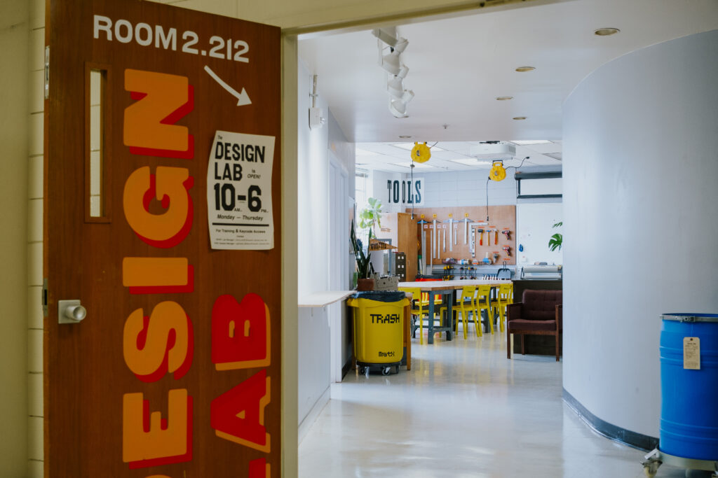 Photo from outside the Design Lab at The University of Texas at Austin. The entrance door is open, and behind it is a hallway leading to the main working area in the space. There are tables and chairs, and on the walls there are design tools like rules and blades hung on a peg board.