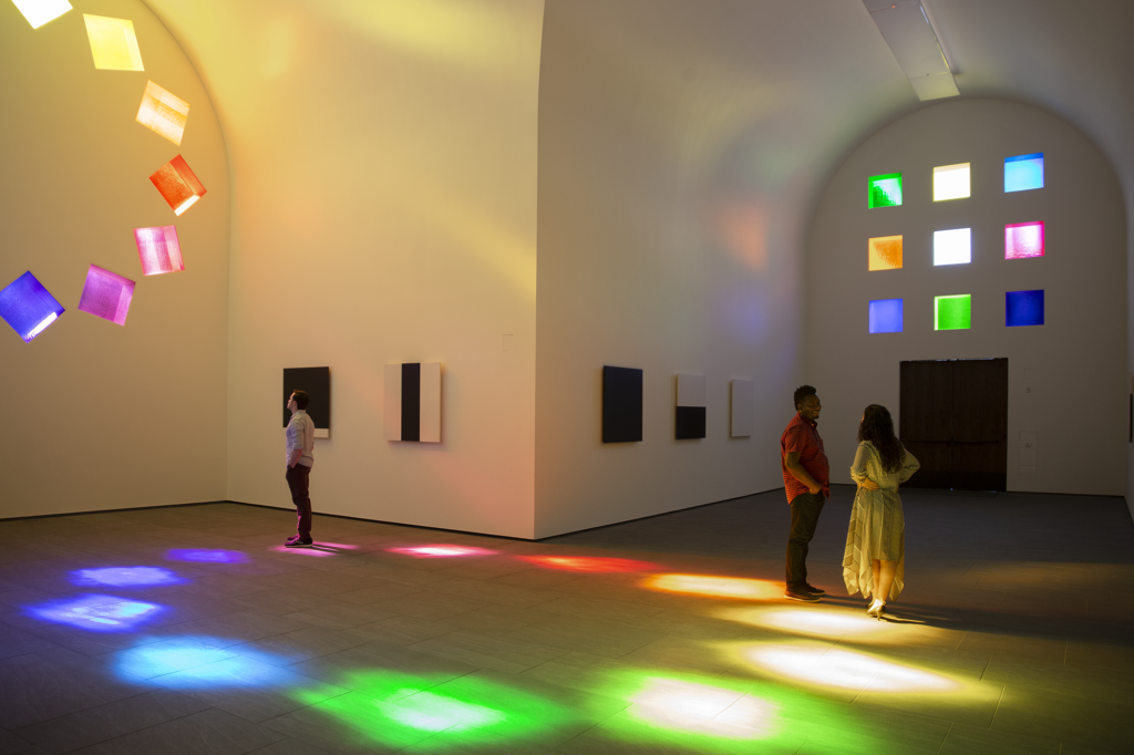 Three patrons stand inside Ellsworth Kelly's large scale art piece titled "Austin." This piece is a full sized building, with two collections of stained glass on two of the walls. The stained glass cast colored shadows onto the floors and walls. There are 5 pieces of art on canvases hanging on the walls as well. Those pieces are black and white stripes assembled in different orders and sizes.