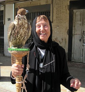 Denise Schmandt-Besserat, a light-skinned person in black clothing with their head covered, stands in front of a wall holding a wooden perch on which a hooded raptor sits.