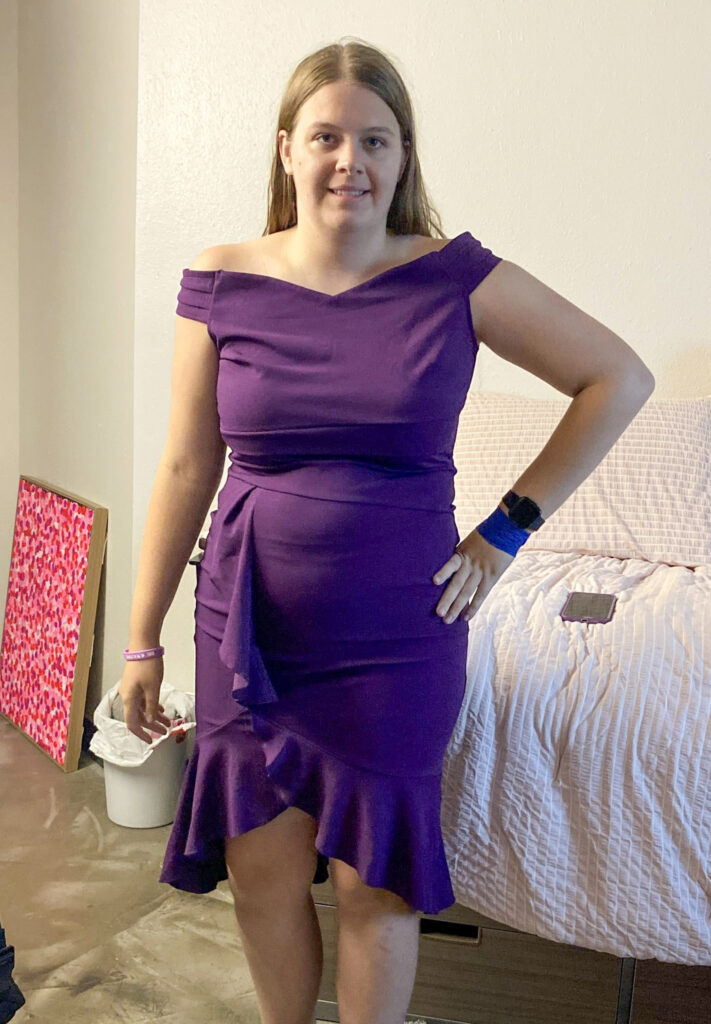 Female student in her dorm wearing a purple cocktail dress with her hand on her hip before going to a dance