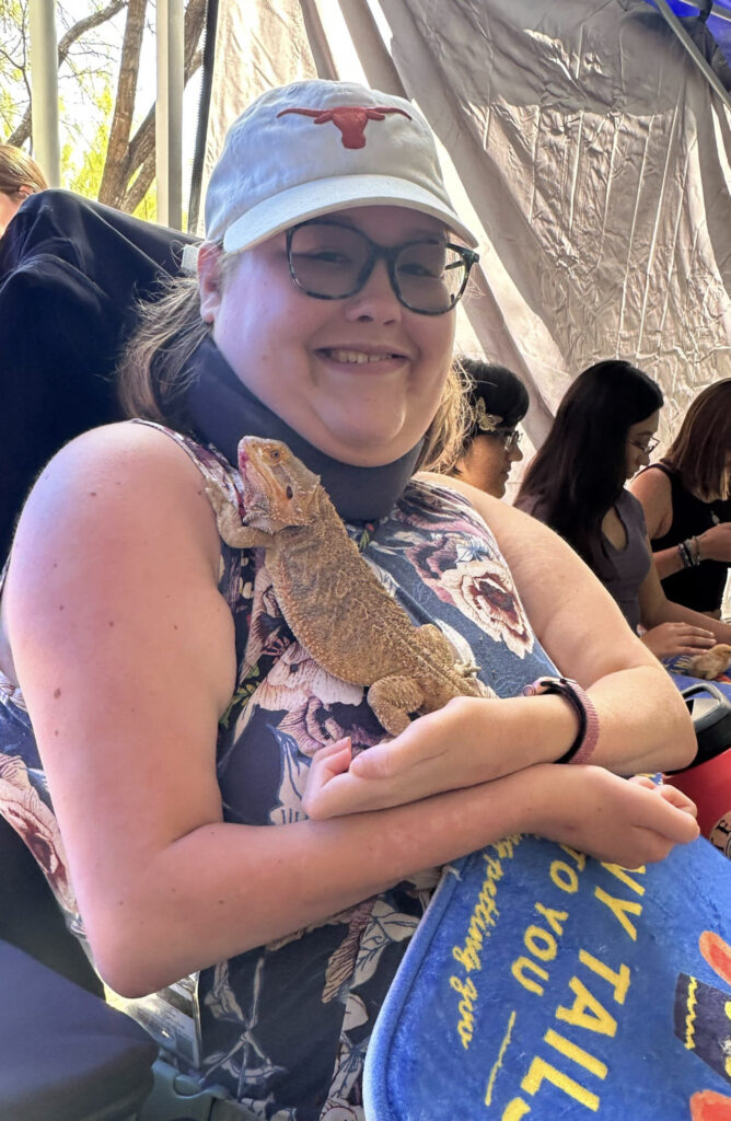 Female student in a UT baseball cap holds a 10 inch tan lizard on her shoulder and chest as part of a visiting petting zoo