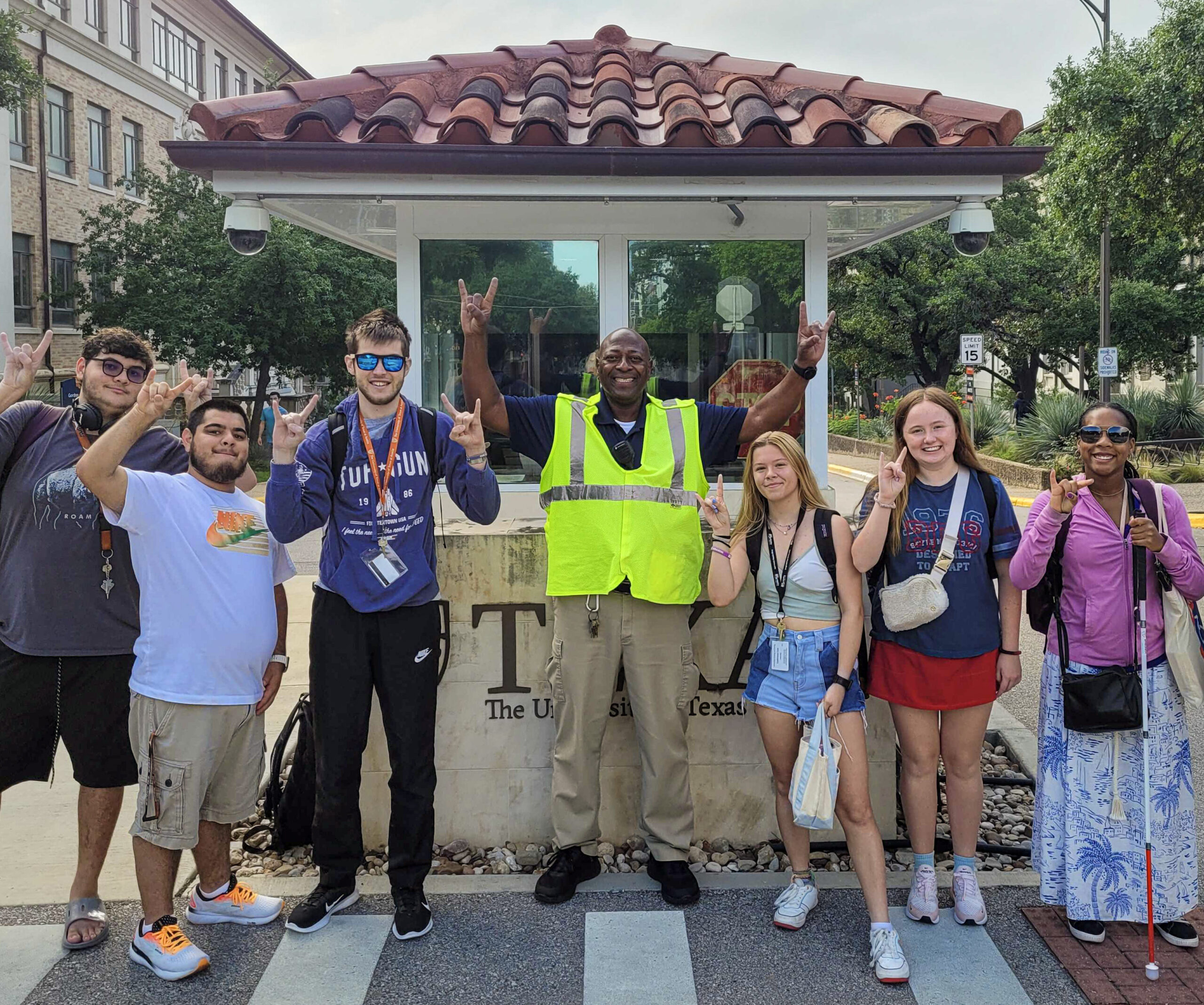 Six male and female students giving the "hook 'em" hand sign stand outside a parking kiosk on campus with a UT employee in a yellow safety vest 