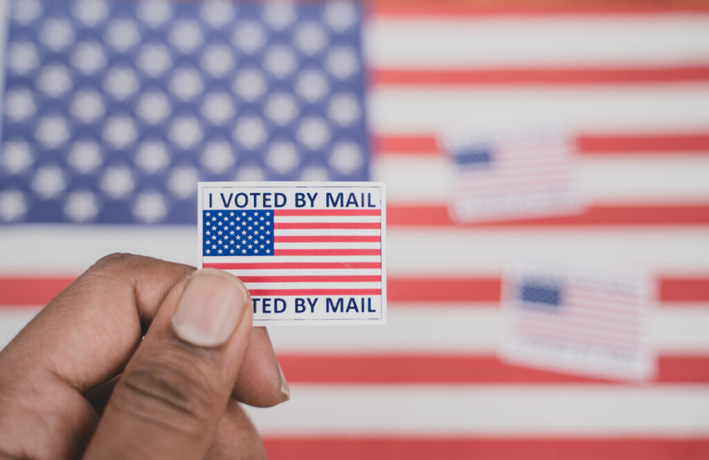 an image of a hand holding a sticker that reads "I voted by mail" with an American flog on it. 