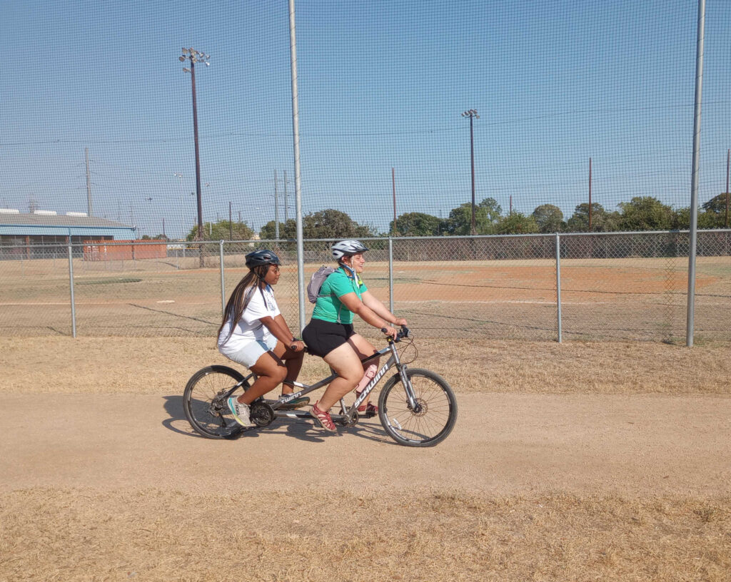 E4Texas female student rides on the back of an adaptive bike to support another female while she rides outside on a tan dirt track 