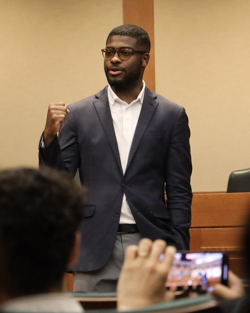 Isaiah Martin, African American Male in a navy sport coat, holds fist in front of his chest as he speaks to a group of students at the State Capitol