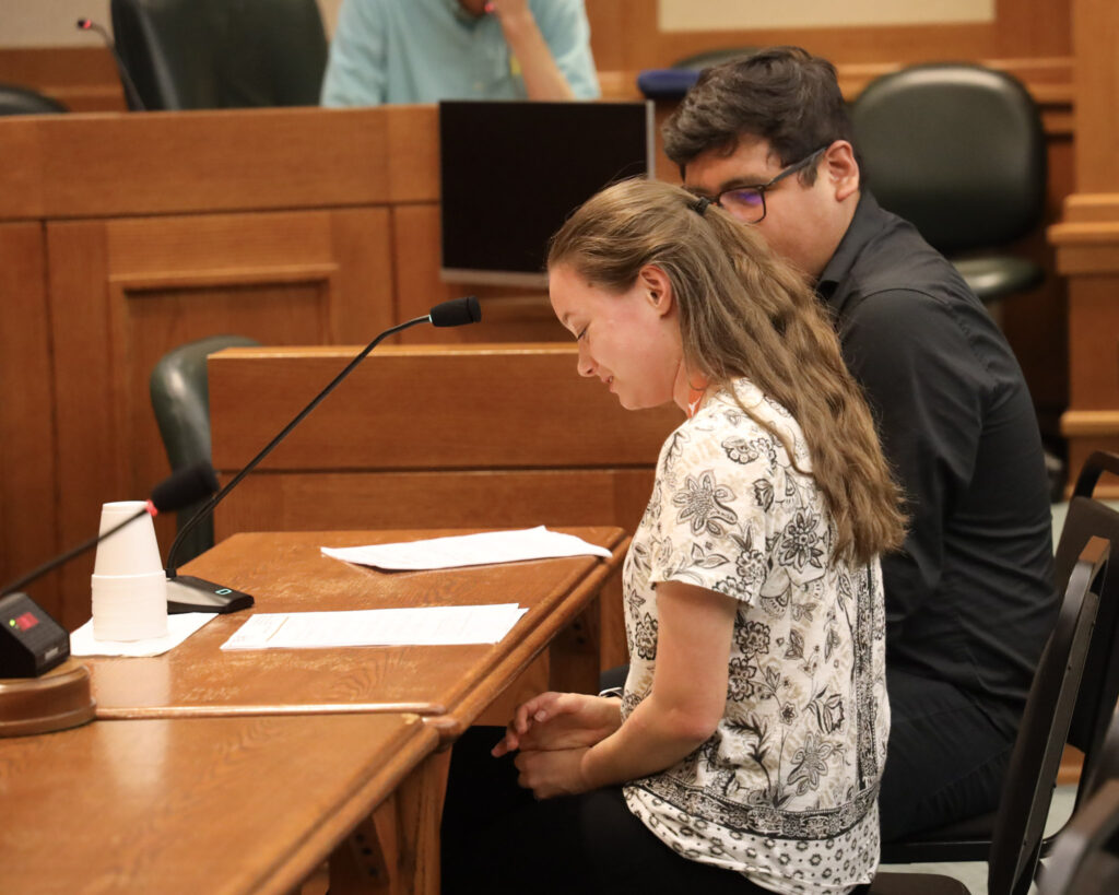 White female student sits with Hispanic male student at a table with a microphone as the female student prepares to speak in front of a panel