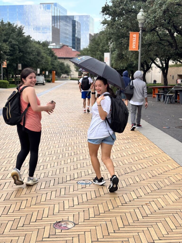 Two white college-aged women walking on a yellow brick paved road with an umbrella, smiling and one is waving