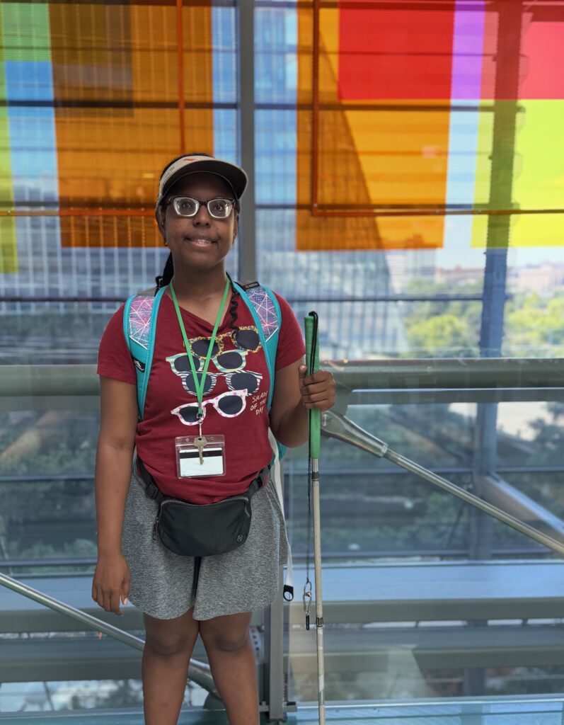 African American female student wearing glasses and a visor stands in front of an orange and yellow art installation/window at the Austin Public Library