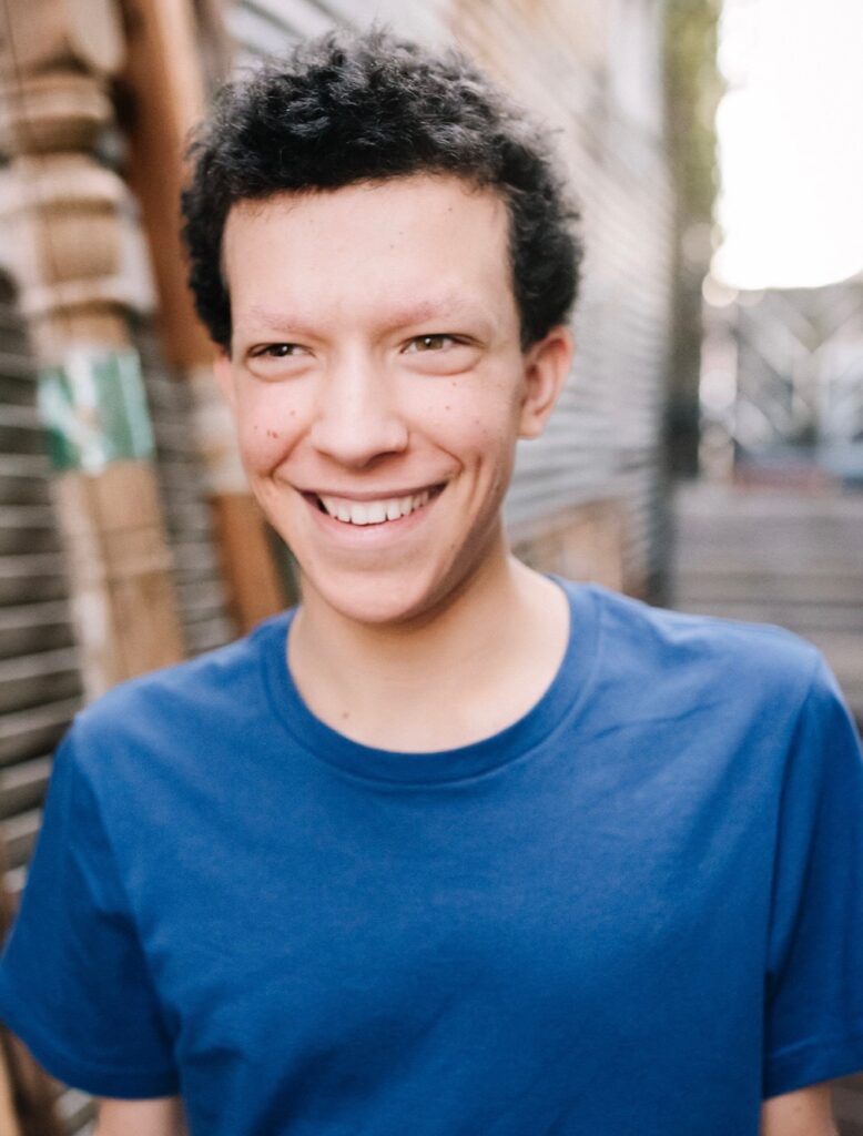 Eli, a young man with short curly dark hair, smiling warmly at the camera. He's wearing a blue t-shirt and standing in front of a corrugated metal wall in a sunlit alley.