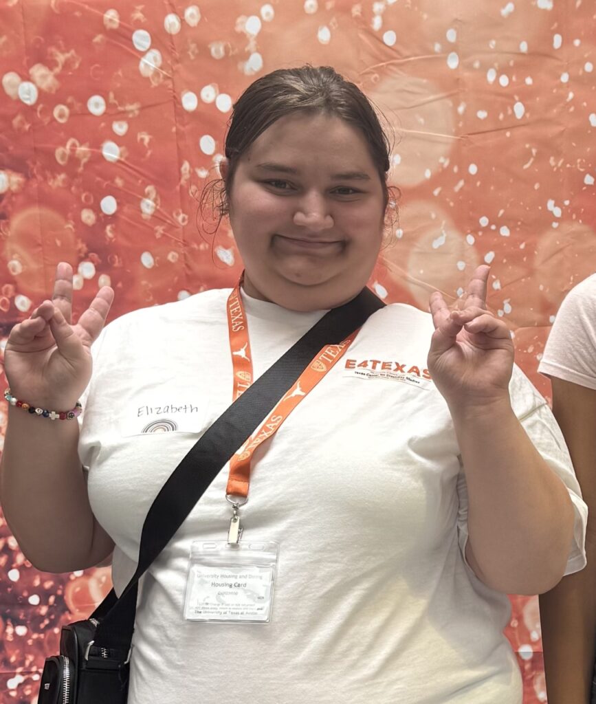 Elizabeth, wearing a white E4Texas t-shirt with an orange lanyard and housing ID badge, smiling and holding up two Hook 'em Horns hand signs in front of an orange and white patterned backdrop.