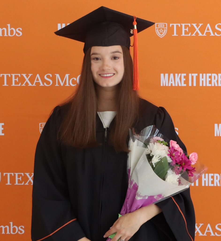 Katelyn smiling in a black graduation cap and gown with an orange tassel, holding a bouquet of white and pink flowers, standing in front of an orange Texas McCombs step-and-repeat backdrop.
