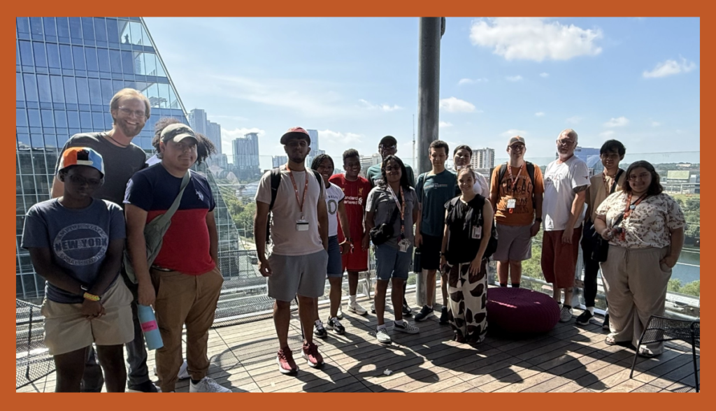 A group of about 14 students and staff posing together on an outdoor rooftop terrace with downtown Austin high-rises in the background under a sunny sky.