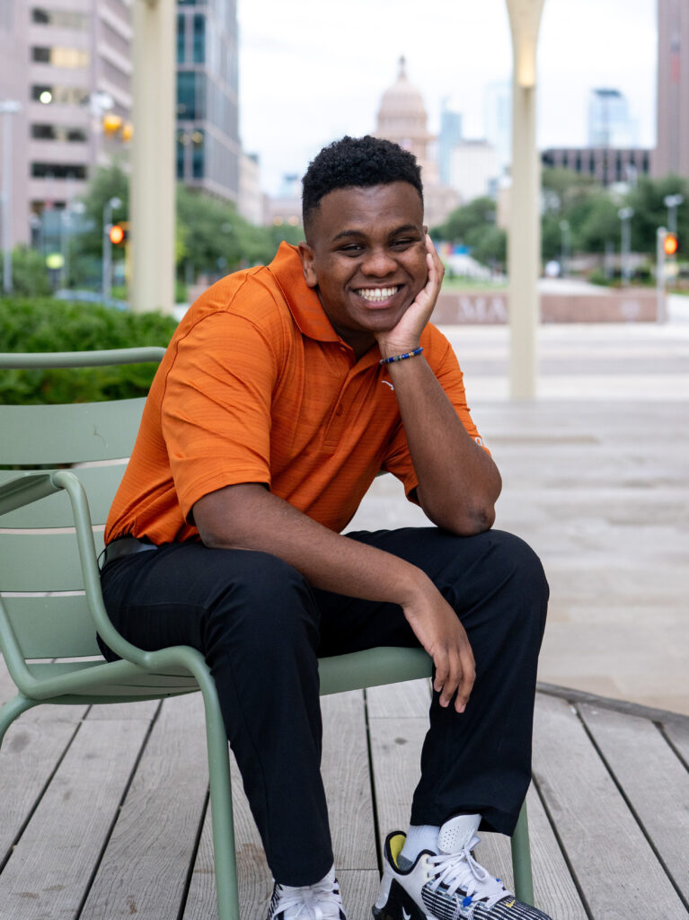 Zane, wearing an orange polo shirt and black pants, sitting on an outdoor bench with his chin resting on his hand, laughing. The Texas State Capitol building is visible in the background.