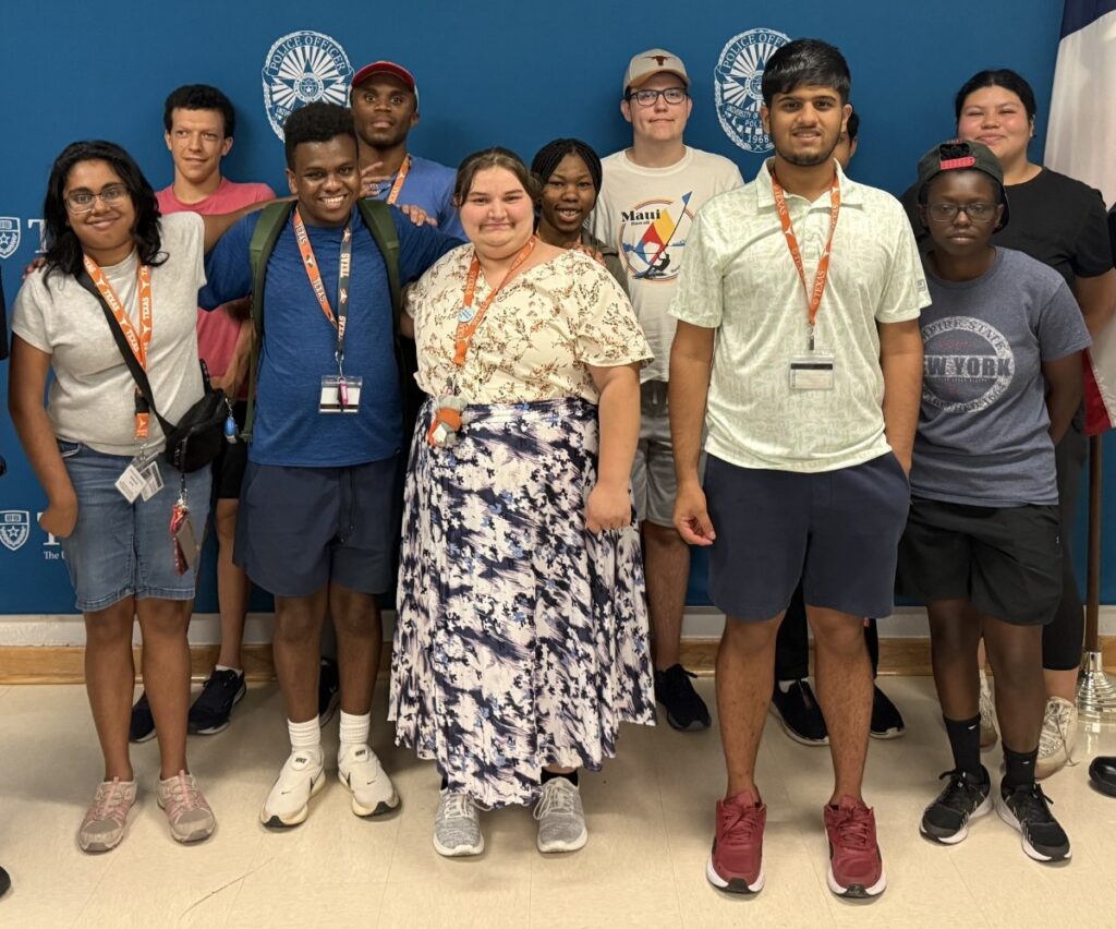 A group of students pose in front of a blue UT Austin PD backdrop with American and Texas flags, several making "hook 'em horns" hand signs.