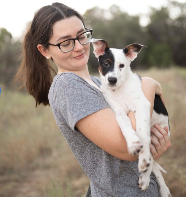 A woman with glasses and dark hair smiles gently while holding a black-and-white puppy with large upright ears, standing outdoors in a grassy field.