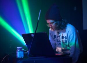 An Arts and Entertainment Technologies student works on a lighting design project on his computer, with green and blue lights shining behind heim.