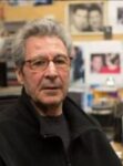 Photo of Yacov Sharir looking into the camera. He has gray hair and glasses and is wearing a dark gray shirt and black jacket.