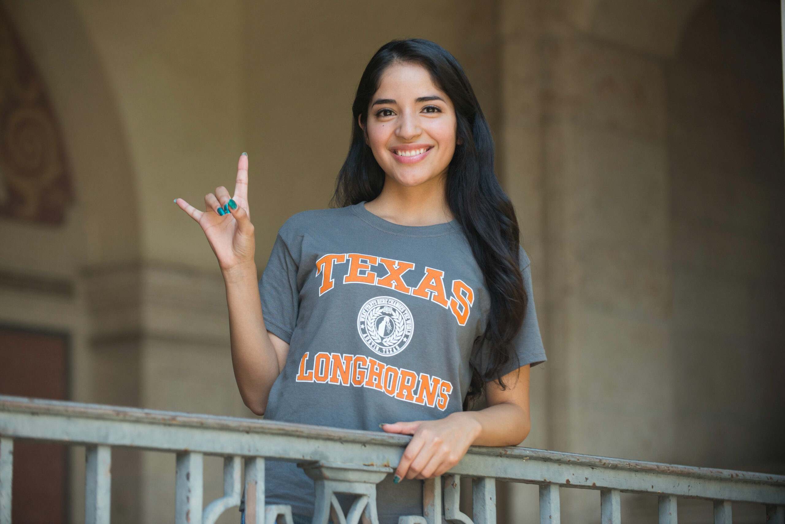 A young woman wearing a gray 'Texas Longhorns' T-shirt with an orange logo. She is smiling and holding up the Texas Longhorns hand sign