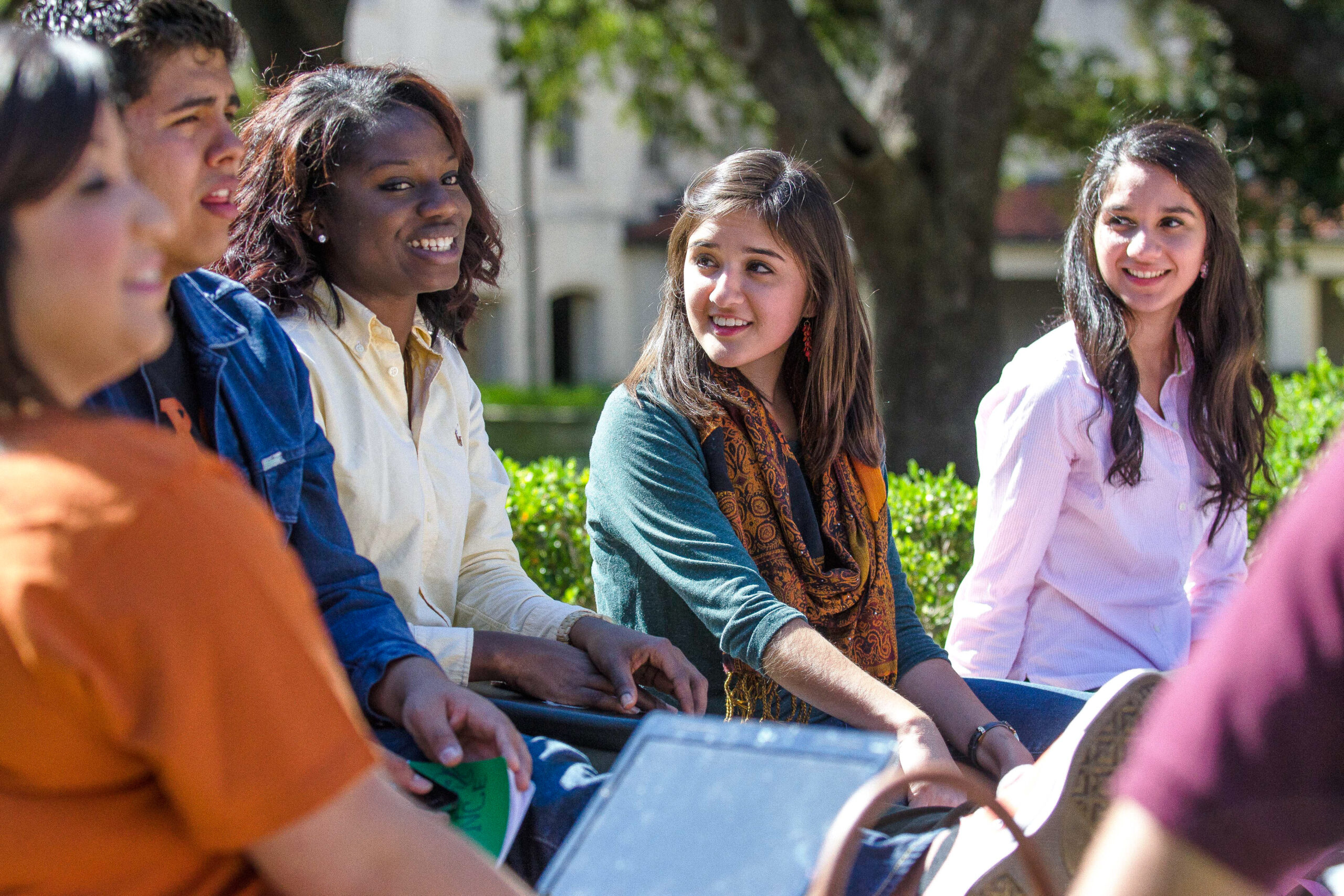 A group of five students sitting together outdoors, smiling and chatting. The students are sitting on a bench surrounded by greenery, and one student is holding a tablet. They appear to be having a lively conversation on a sunny day.