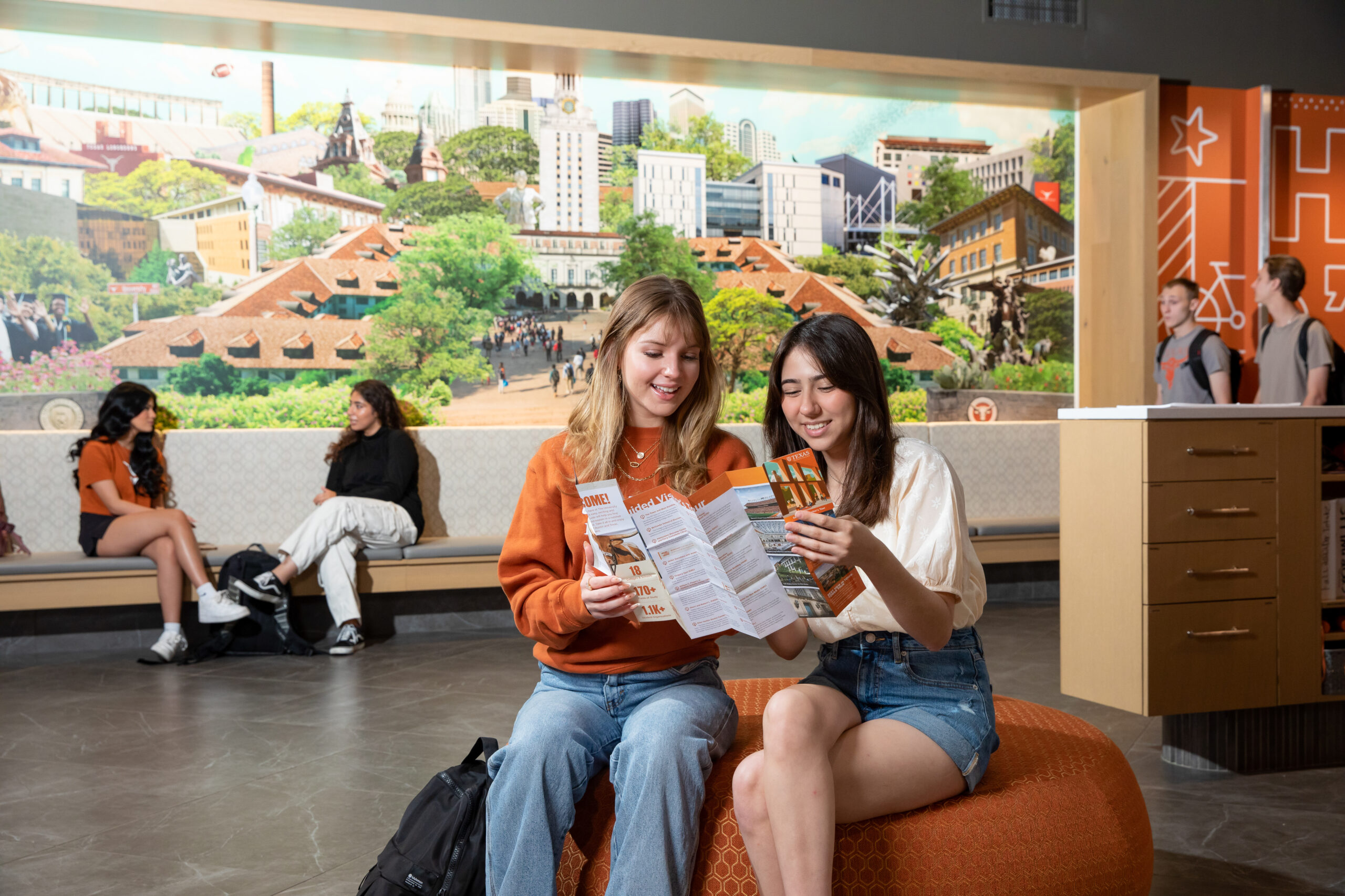 Two students sit on an ottoman in a bright room, reading through brochures. A mural of the University of Texas campus is in the background, with other students sitting and conversing in the background.