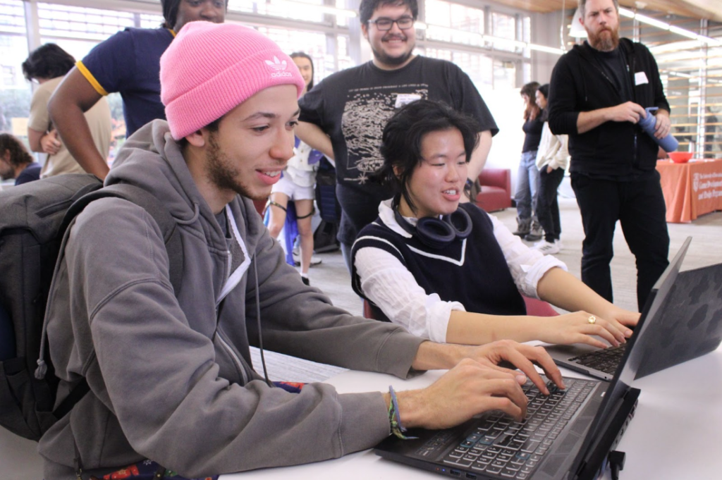 A group of students engaging in a collaborative activity at a workshop. Two students are seated at a table, smiling and typing on laptops, including one wearing a pink beanie and another with headphones around their neck. Other participants and an instructor are visible in the background, observing and interacting in a brightly lit, open space.