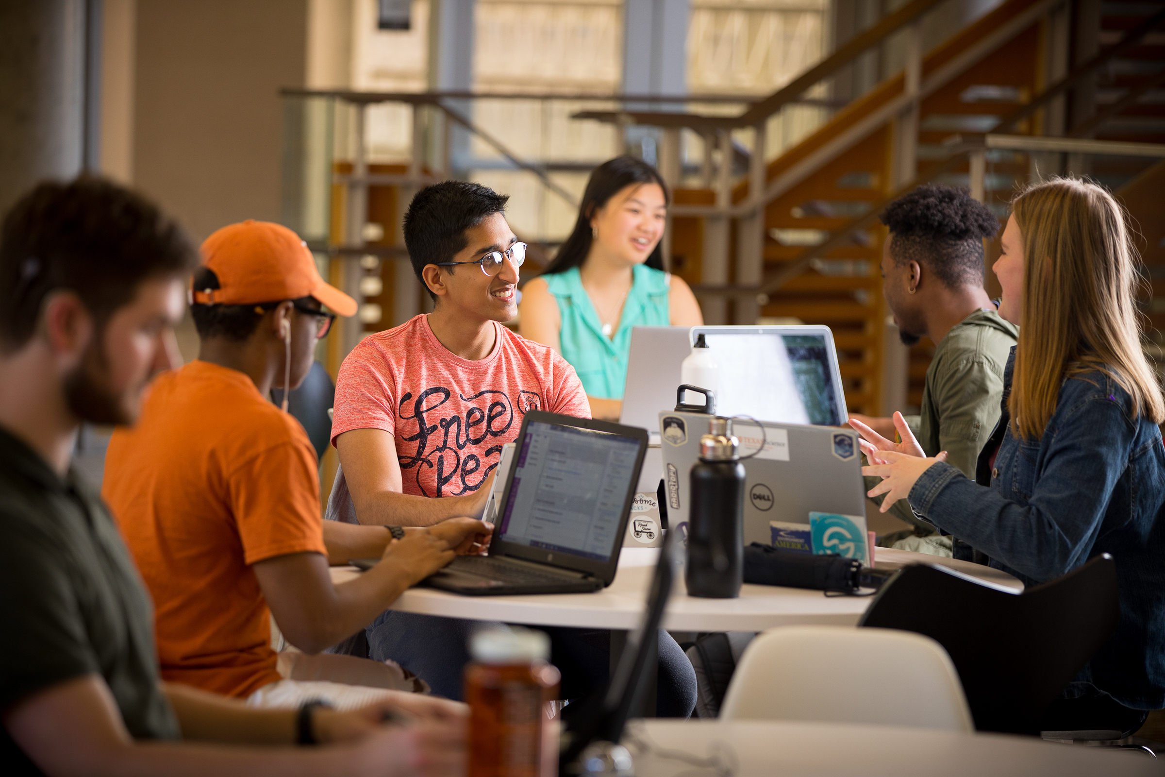 Several students are seated around a table in a bright, spacious room, working on laptops and discussing. They appear to be collaborating on a project, with a large staircase and open seating visible in the background.