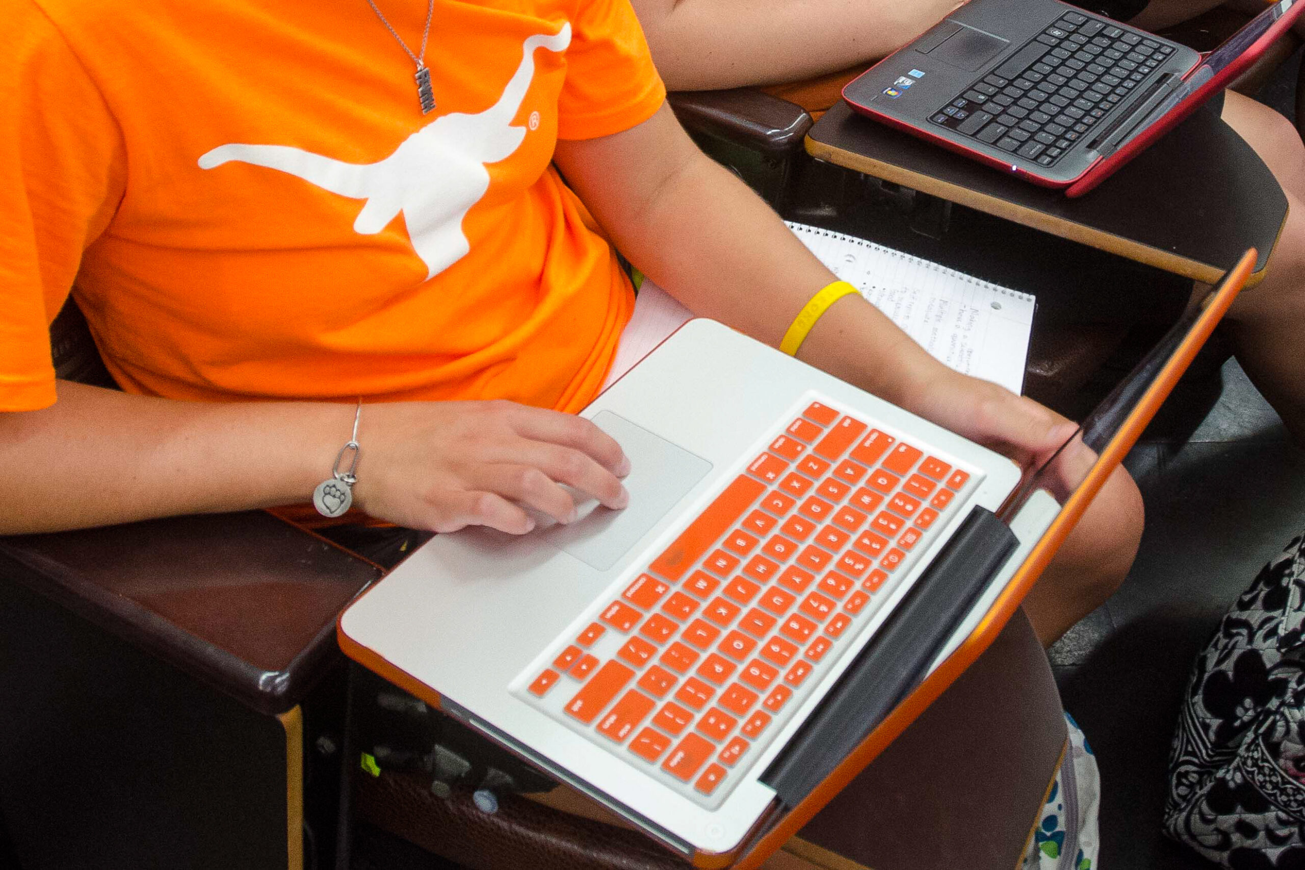A student wearing glasses works on a laptop while referencing notes in a sketchbook, with an architectural model in the foreground. Another student, wearing a backwards cap, leans in to observe. The classroom is filled with other students working on computers and models, with large windows providing natural light.