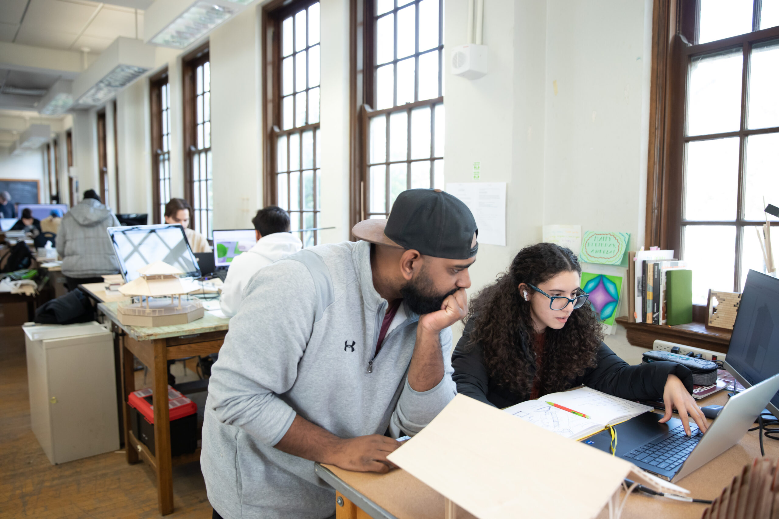 A student wearing glasses works on a laptop while referencing notes in a sketchbook, with an architectural model in the foreground. Another student, wearing a backwards cap, leans in to observe. The classroom is filled with other students working on computers and models, with large windows providing natural light.