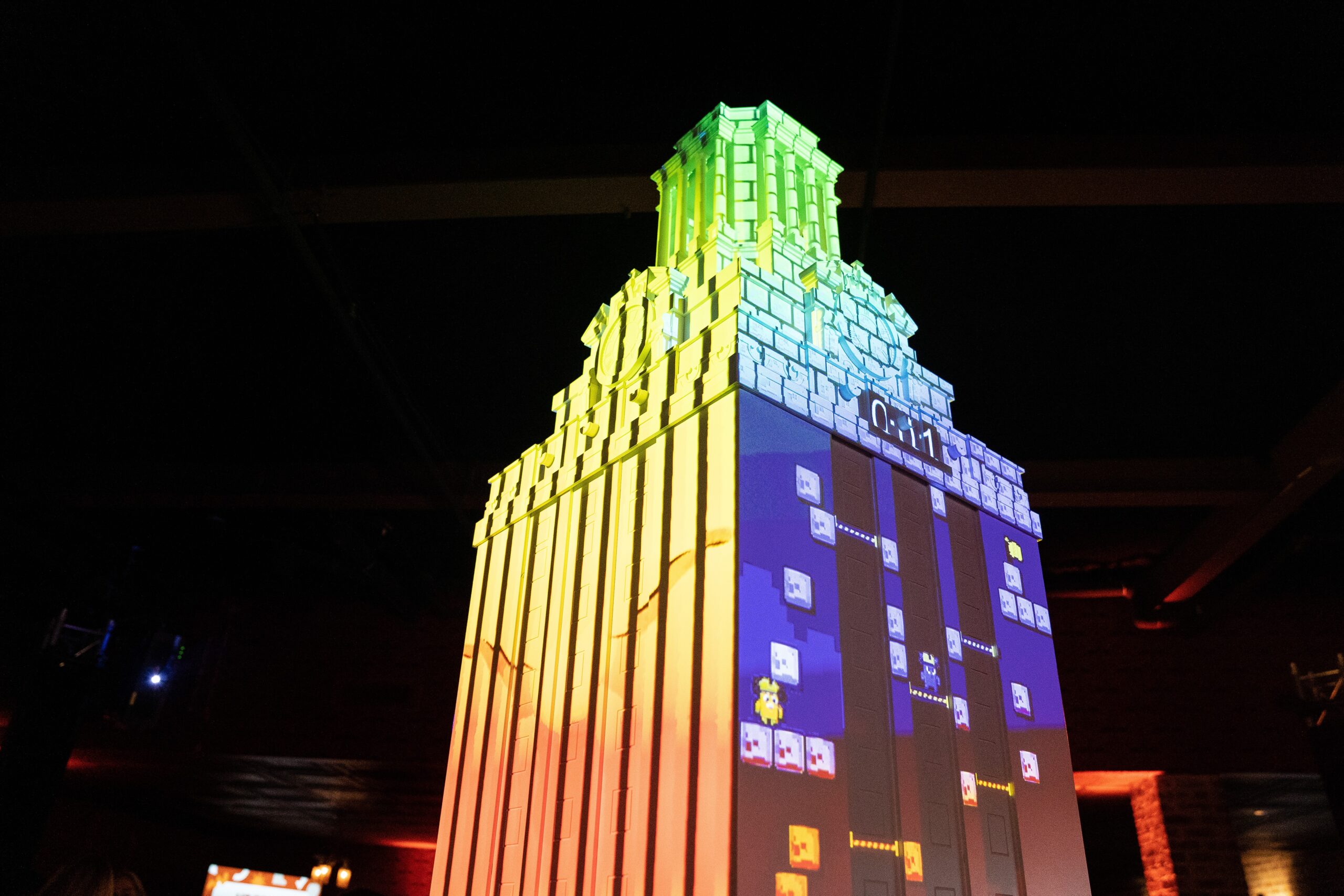 A large 3D projection of the University of Texas tower, illuminated with rainbow colors, showcases a video game display. The tower projection includes blocks and game characters, creating a vibrant gaming experience.