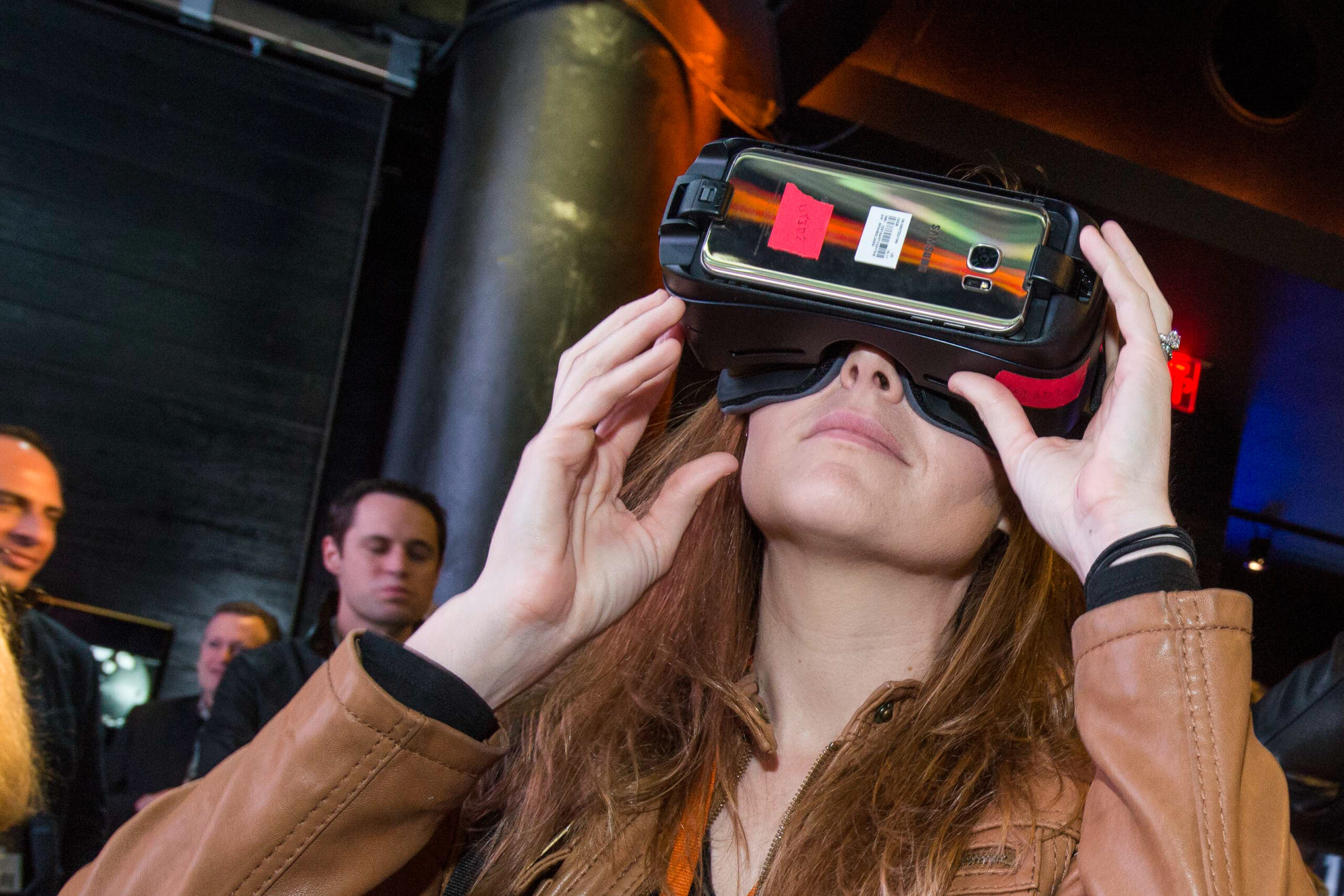 Two participants wearing virtual reality headsets, immersed in an experience. The person on the left, a woman in a brown jacket, adjusts her headset, while the person on the right smiles while looking through the headset. They are in a dimly lit room with several other onlookers in the background.