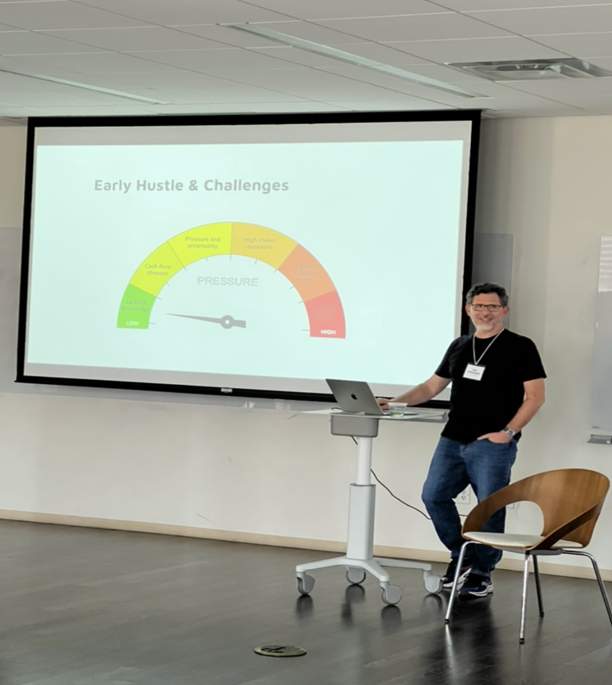 A smiling speaker stands next to a rolling podium with a laptop, presenting a slide titled “Early Hustle & Challenges.” The slide displays a semicircular gauge labeled “PRESSURE” ranging from low (green) to high (red), with indicators for challenges like cash flow stress, pressure and uncertainty, and high-stakes decisions.