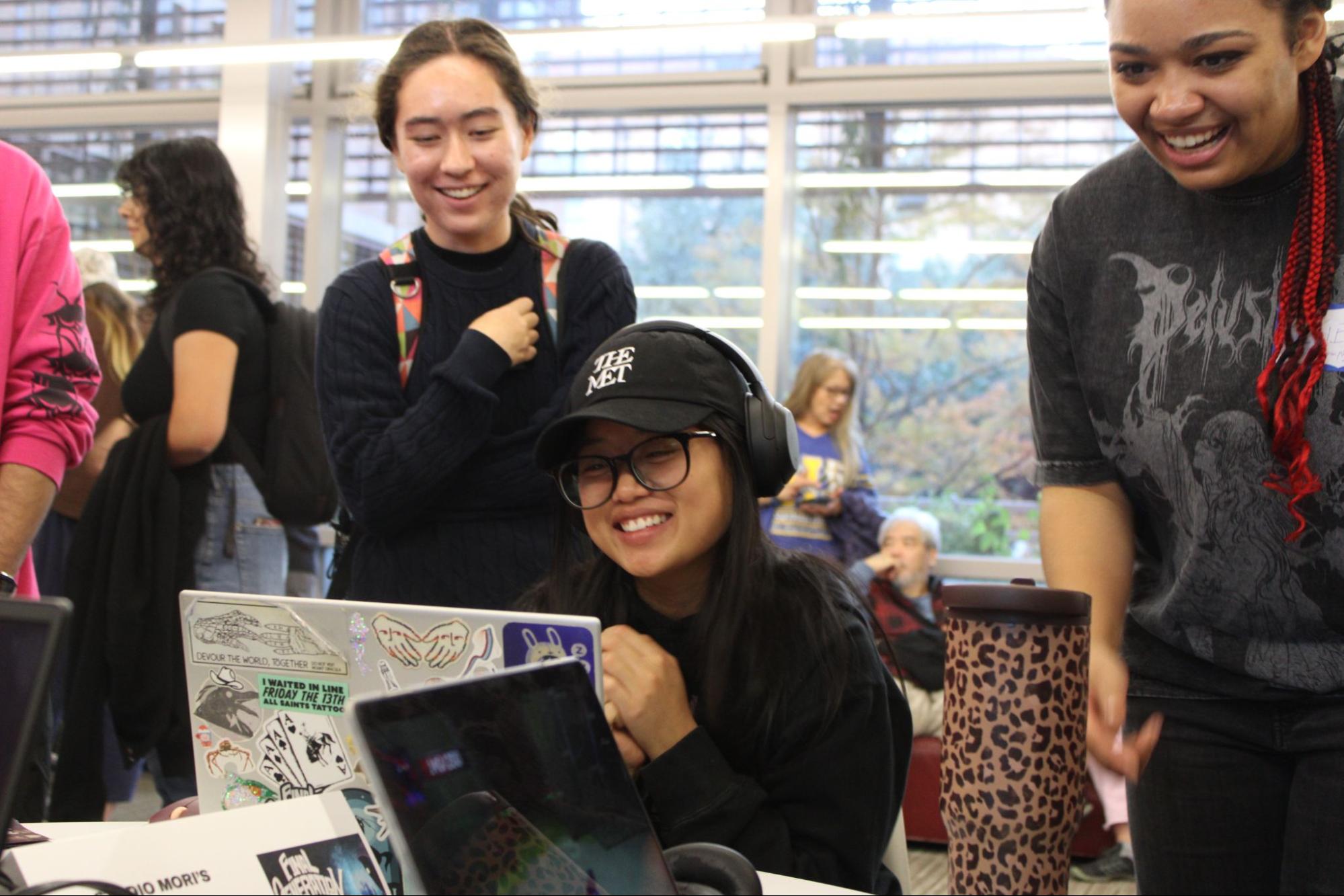 Three students gather around a laptop covered in playful stickers during Digital Demo Day. One student smiles brightly while wearing headphones and a cap that reads “THE MET.” The group is engaged and joyful, highlighting the collaborative and celebratory spirit of the event.
