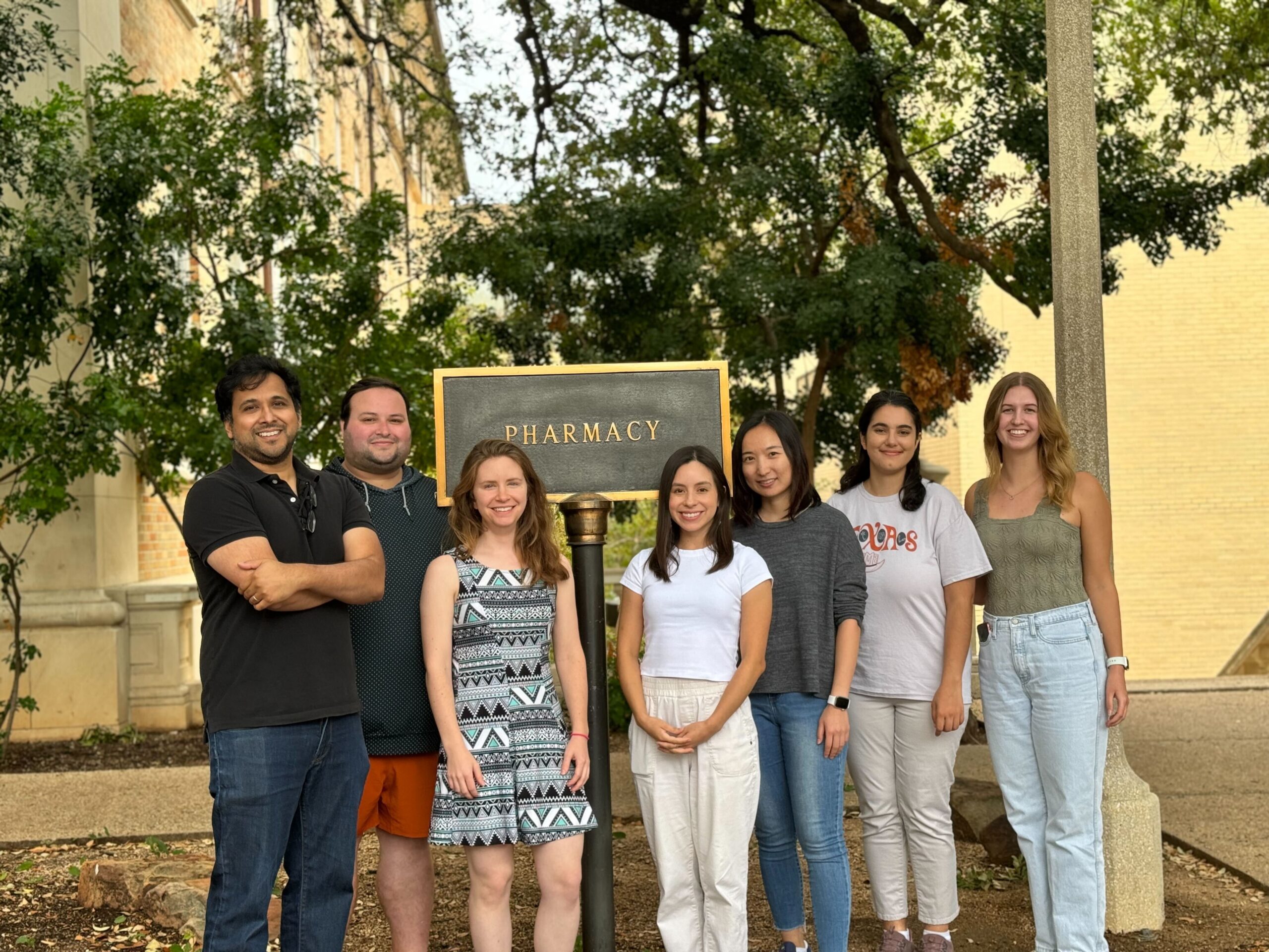 Dr. Ghosh and six lab members smiling and standing in front of the Pharmacy building sign