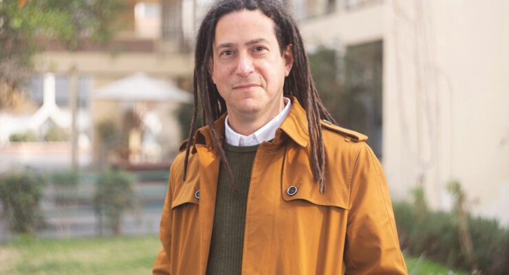 A white man with brown dreadlocks looks at the camera. He is wearing a gold-orange colored jacket, an olive green sweater, and his shirt has a white collar. He is outdoors in front of a building with some grass and a patio visible.