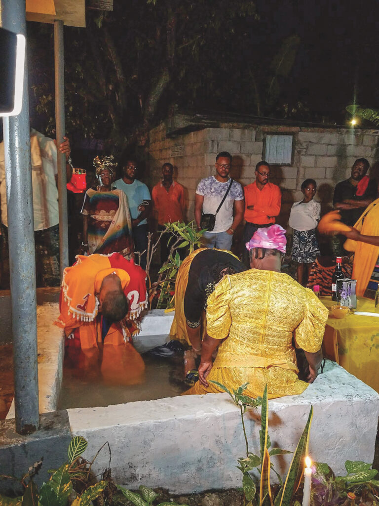 A group of Black men, women, and children are gathered for a ritual. Three are dressed ornately and are in a small concrete pool. Others look on in front of a simple one-story cinderblock building. One woman has a metallic silver head wrap. It is nighttime. In the foreground, a candle is lit in a garden bed.