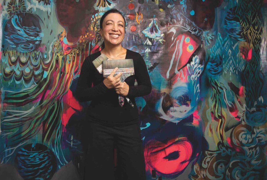 A broadly smiling woman dressed in black holds two large paint brushes and stands in front of a large multicolored abstract painting that contains images of water. The painting is taller than the woman and takes up the entire frame of the photo.