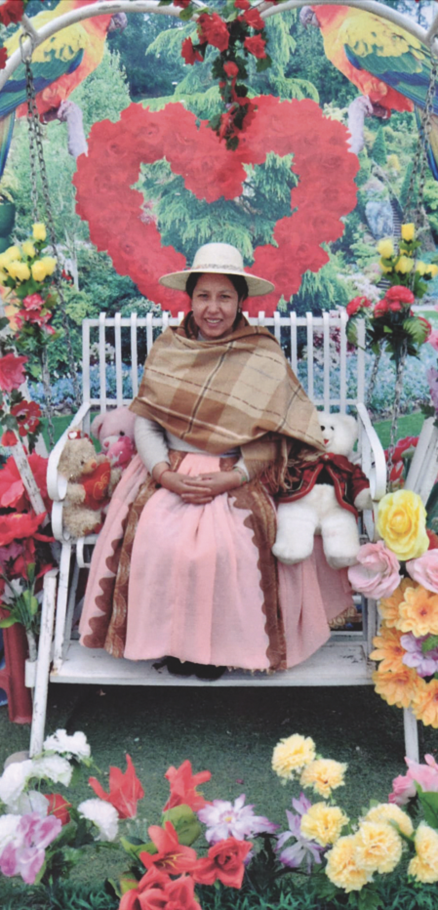 A woman dressed in traditional Andean polleras sits on a white love seta surrounded by three stuffed teddy bears. There are flowers in various places around her, and there is a wall in back with a photograph of a heart made of red roses and large colorful parrots, almost out of the frame. Centered, she smiles. She is wearing an off-white straw hat, a shawl in brown plaid draped across her chest, and a pale pink full-length skirt with brown accents sewn on vertically in two places. Her hands are folded. Her hair is black and gathered behind.